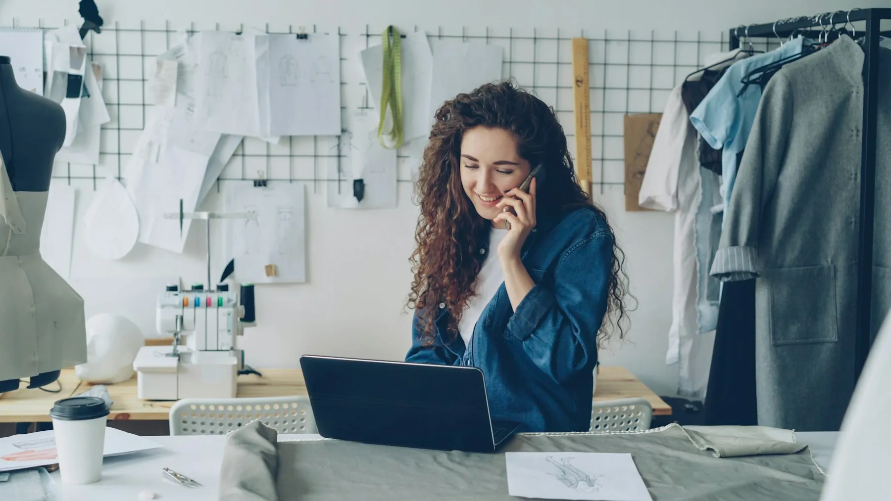 Diseñadora de moda sonriendo mientras habla por teléfono frente a su laptop en su taller